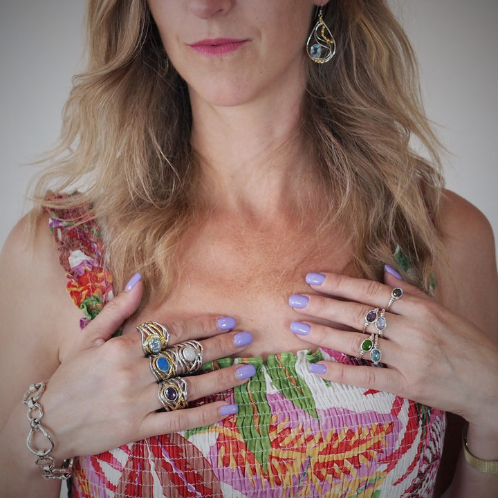 A woman with lavender-painted nails wears multiple Gallardo & Blaine Designs' Mayhem Rings in various gemstones. She has long, wavy blonde hair and dons a colorful, patterned top with thin straps. She also wears a bracelet and dangling earrings that match her rings.