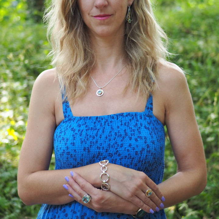 A woman with wavy blonde hair stands outdoors wearing a blue patterned sundress. She has a necklace, earrings, and a statement Gallardo & Blaine Designs Sea Honey Bracelet Silver with large, thick silver links. Her nails are painted lilac, and she has multiple rings on her fingers. The background is lush greenery.