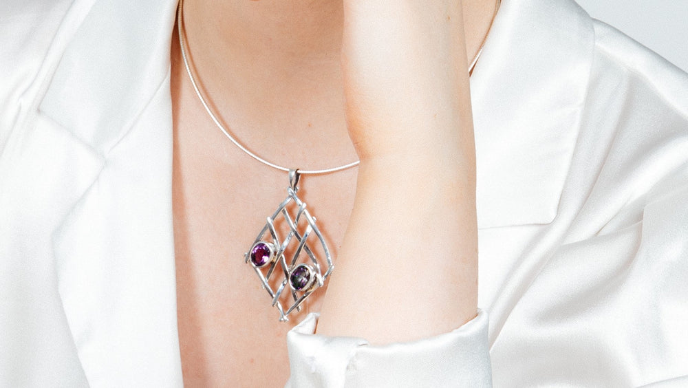 A person in a white satin shirt displays a Gallardo & Blaine Designs sterling silver necklace with a Nettle Pendant featuring gemstone combinations in a lattice pattern, gently touching their neck.