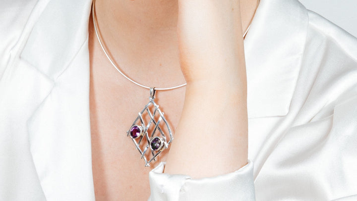 A person in a white satin shirt displays a Gallardo & Blaine Designs sterling silver necklace with a Nettle Pendant featuring gemstone combinations in a lattice pattern, gently touching their neck.