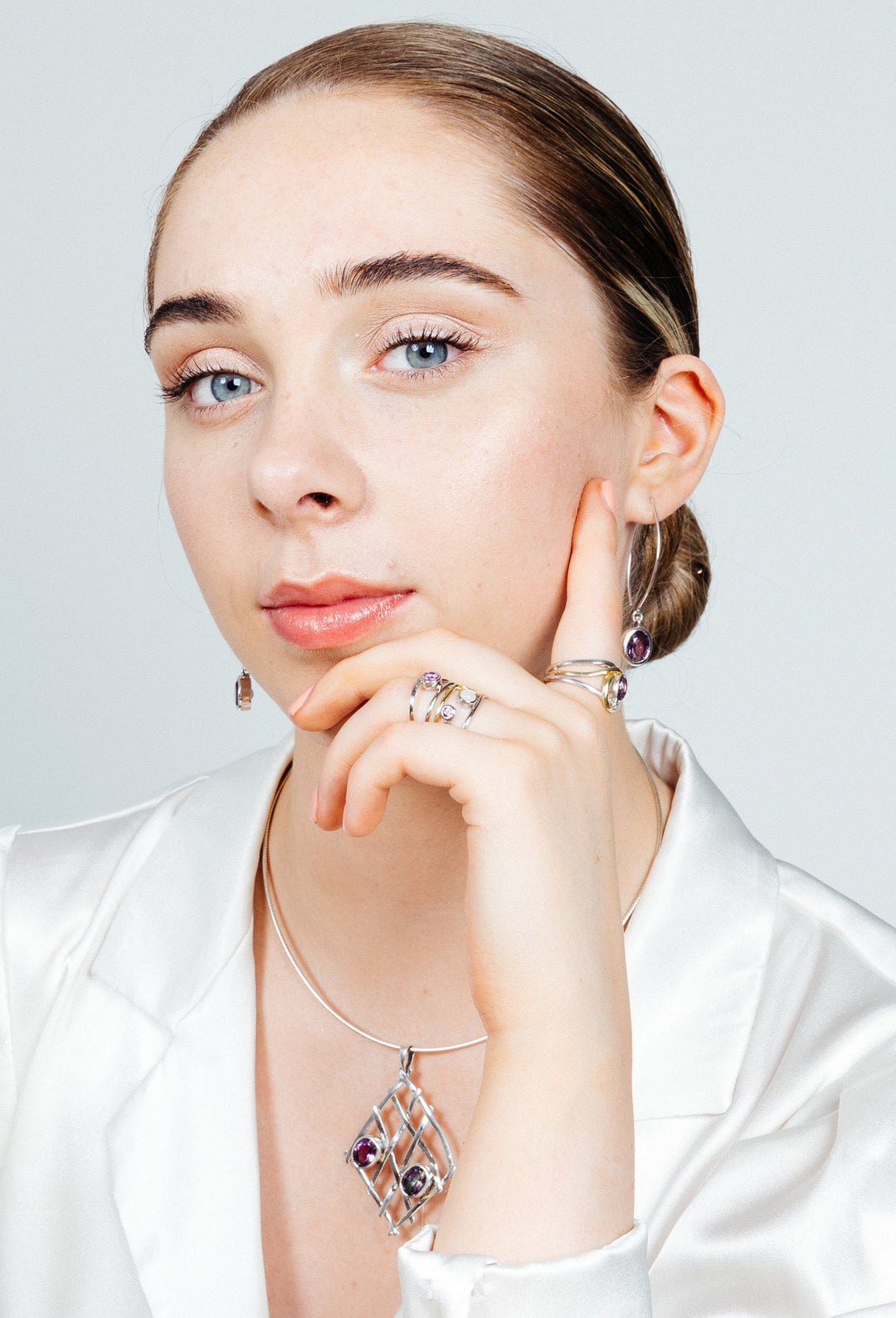 A blue-eyed individual in a white satin top poses with a hand on their chin, showcasing Gallardo & Blaine Designs' Nettle Pendant in various gemstones. They wear matching sterling silver earrings and rings with purple stones against a plain white background.