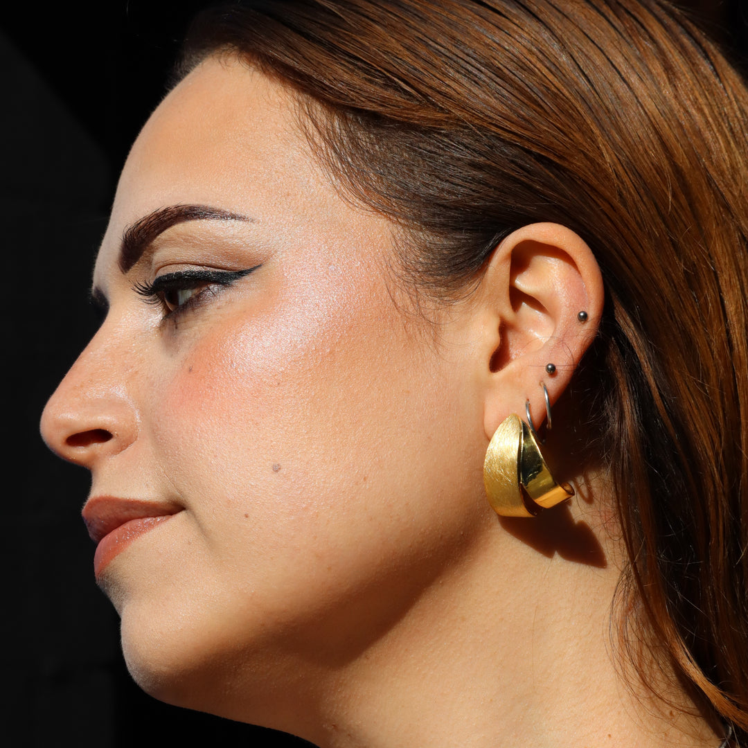 A woman with light brown hair, wearing Gallardo & Blaine Designs Origata Studs and multiple small earrings, is shown in profile with winged eyeliner and natural makeup, highlighted by sunlight.