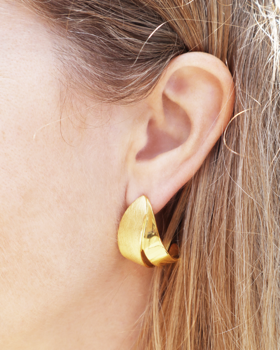 Close-up of a person's ear adorned with Gallardo & Blaine Designs Origata Studs—large, shiny, abstract-shaped gold vermeil earrings—with light brown hair partially visible.