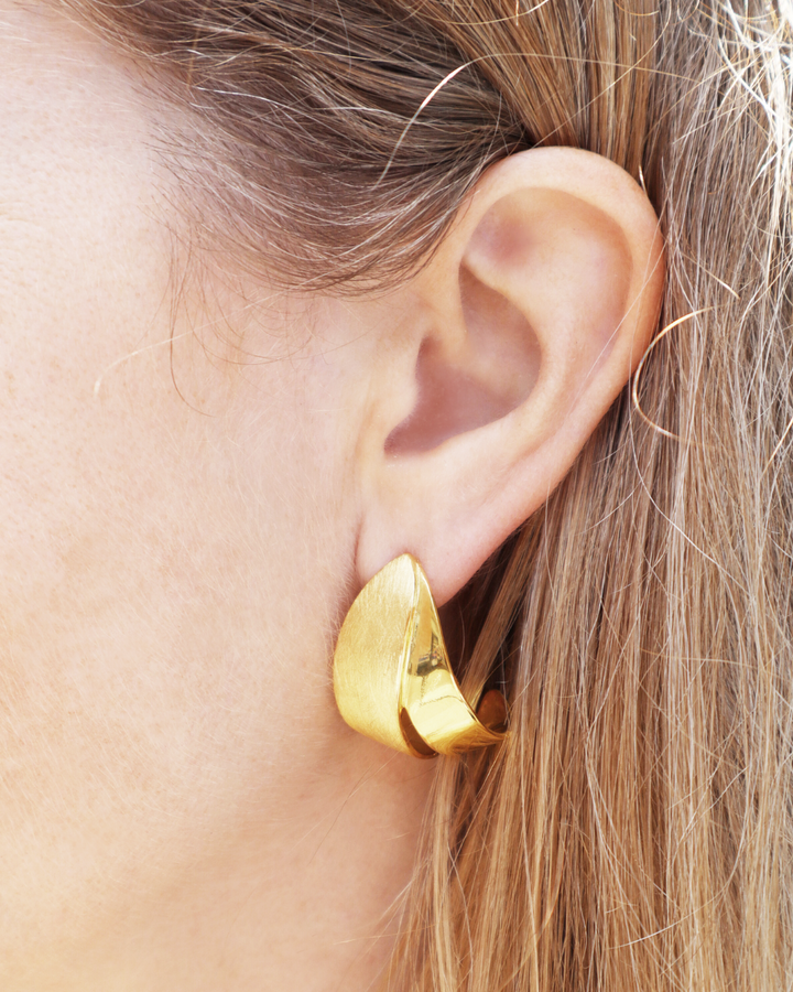Close-up of a person's ear adorned with Gallardo & Blaine Designs Origata Studs—large, shiny, abstract-shaped gold vermeil earrings—with light brown hair partially visible.