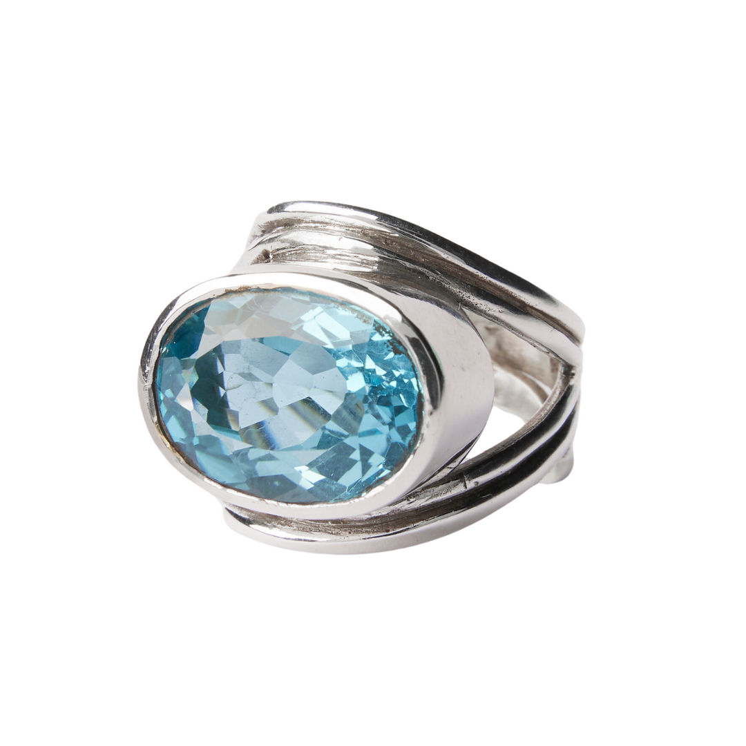 Introducing the Gallardo & Blaine Designs Party Ring in various gemstones, featuring a large oval-cut blue gemstone. The band has a unique, overlapping design with smooth, curved lines that frame the gem, creating an elegant and modern aesthetic. This adjustable statement gemstone ring is both versatile and striking.