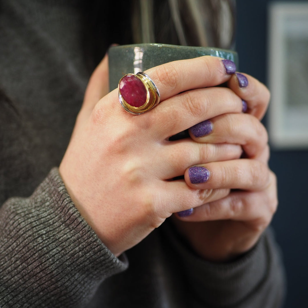 A person with purple glitter nail polish is holding a green ceramic mug with both hands. They are wearing a large, statement Party Ring in various gemstones by Gallardo & Blaine Designs with a gold setting on their left middle finger. The person is dressed in a grey sweater, and the background is blurred.