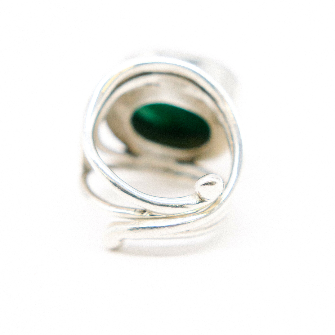 A close-up image of a statement gemstone ring featuring an artistic, twisted design. The adjustable Party Ring in various gemstones by Gallardo & Blaine Designs has an oval green stone set in the center, partially enveloped by the sterling silver bands. The background is a plain white, highlighting the intricate details of the ring.