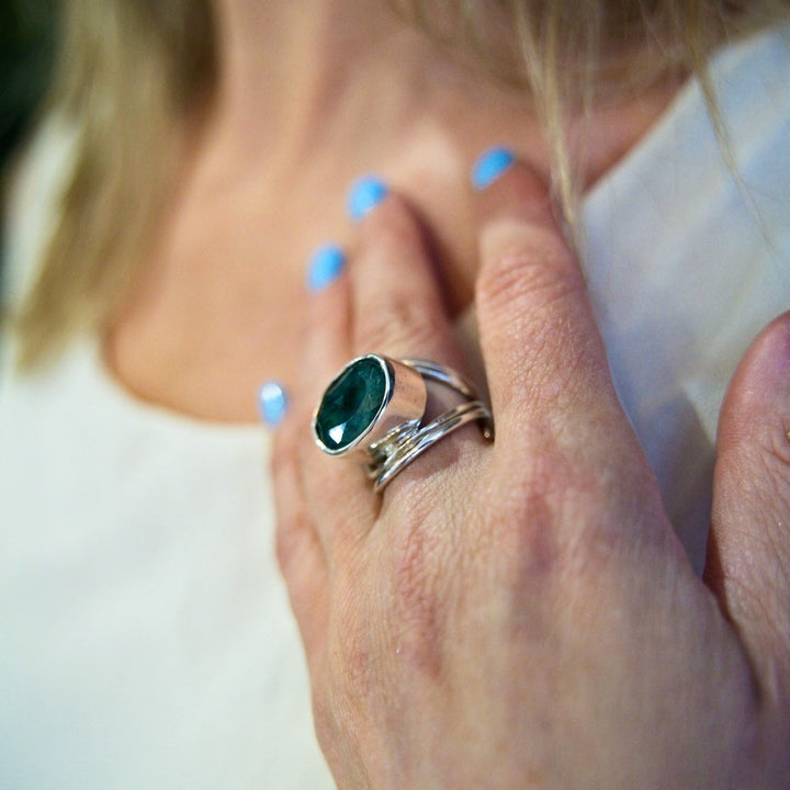 A close-up of a hand adorned with sterling silver and gold rings, including a statement gemstone ring featuring Gallardo & Blaine Designs' Party Ring in various gemstones. The hand, with light blue nail polish, rests against a white top. Blonde hair and a bit of shoulder are visible in the background.