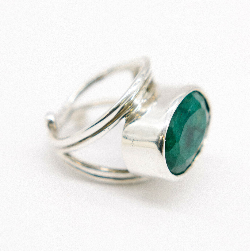 A Party Ring in various gemstones by Gallardo & Blaine Designs featuring a large, oval-shaped green gemstone. The band has a unique, intertwined design in sterling silver and gold, giving the adjustable ring an elegant appearance. The faceted statement gemstone catches light beautifully, adding to the ring's overall shimmer.