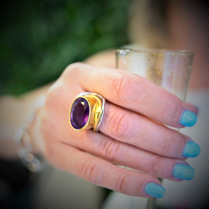 A close-up of a hand with light blue nail polish holding a champagne glass. The person is wearing a large sterling silver & gold Party Ring in various gemstones from Gallardo & Blaine Designs. The background is slightly blurred, with hints of greenery.