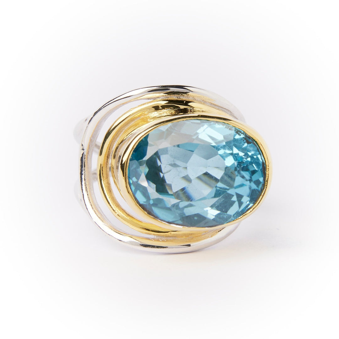 A **Party Ring in various gemstones** featuring a large, round, faceted blue statement gemstone in the center by **Gallardo & Blaine Designs**. The gemstone is encircled by swirling, overlapping gold bands, enhancing the ring's elegant and sophisticated design. The background is a clean, plain white.