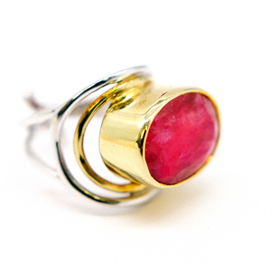 A close-up of an elegant statement gemstone ring featuring a large, oval, pink gem set in a gold bezel. The adjustable ring band is composed of intertwined sterling silver and gold loops, creating a modern and stylish design. The Party Ring in various gemstones by Gallardo & Blaine Designs is displayed against a plain white background.