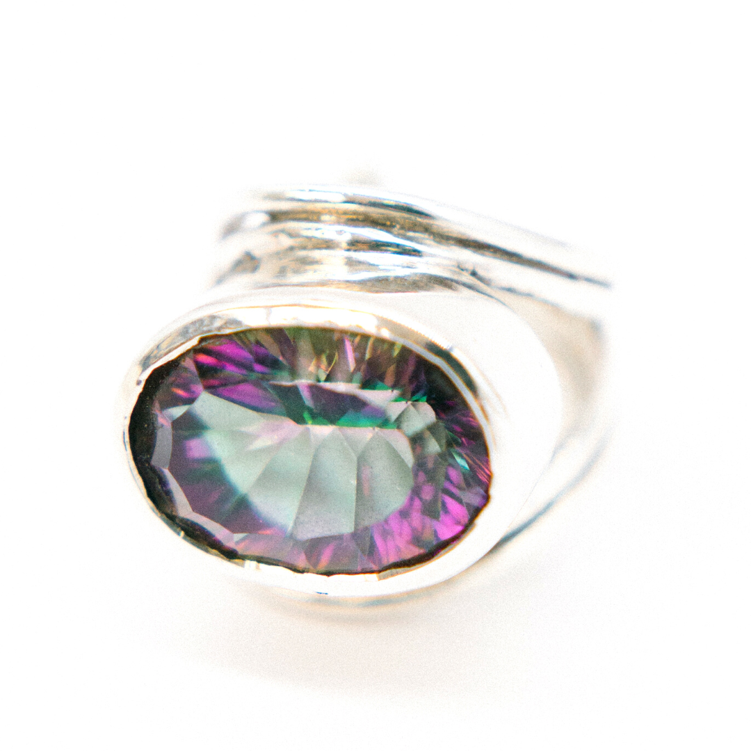 A close-up image of a statement gemstone ring featuring a large, multicolored gemstone with hues of green, purple, and hints of blue, set in a simple bezel setting. The background is plain white, emphasizing the Gallardo & Blaine Designs Party Ring in various gemstones' intricate colors and reflective facets.