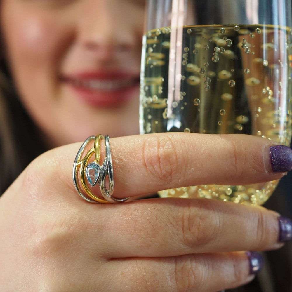 A person with long hair is holding a glass of sparkling wine. They are wearing an elegant, dainty Peacock Ring in various gemstones by Gallardo & Blaine Designs, adjustable in silver and gold, and have purple nail polish. The background is slightly blurred, making the glass and ring the focal points.