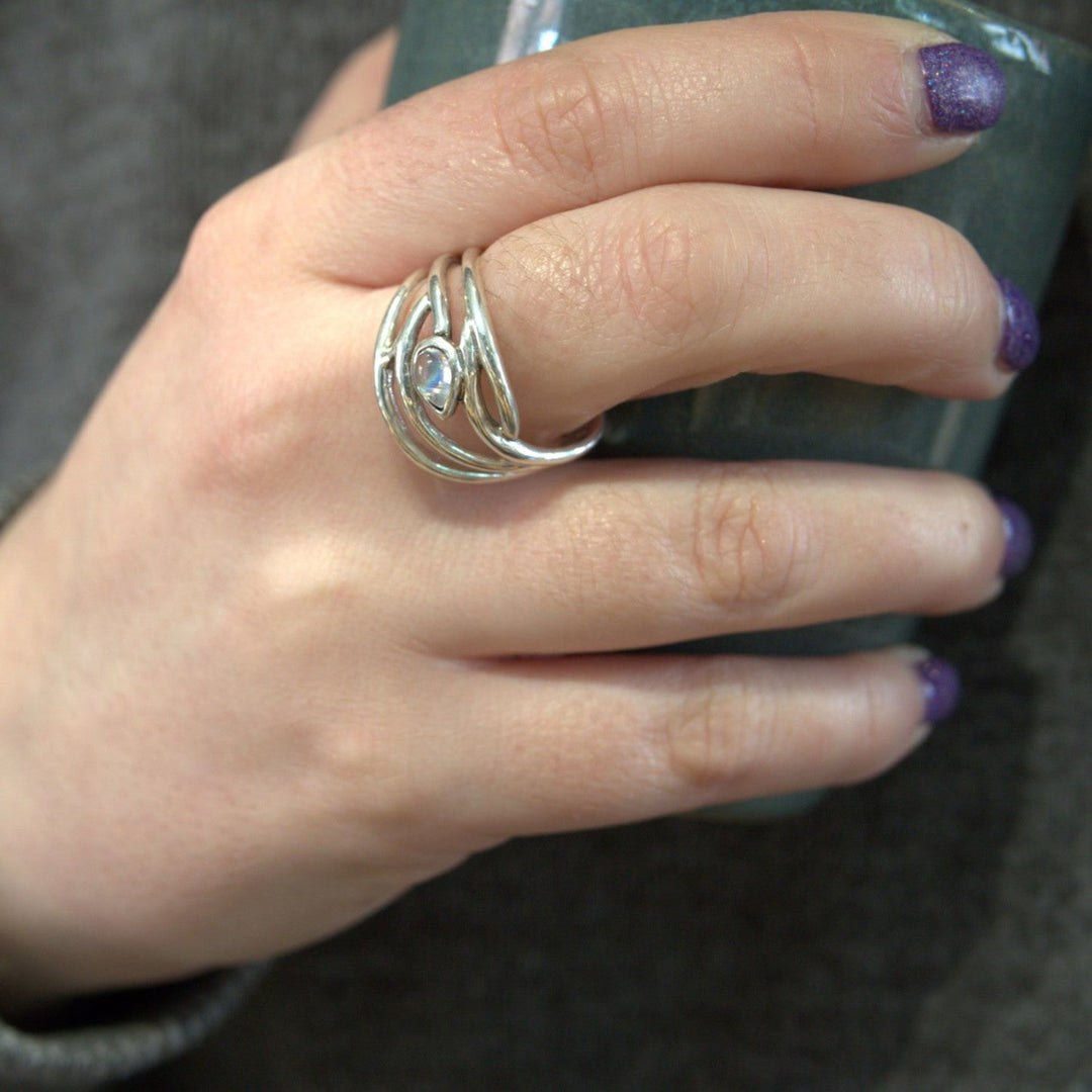 Close-up of a hand holding a greenish-blue ceramic cup. The hand is adorned with an elegant dainty ring featuring an intricate, wavy design and a central gemstone. This beautiful ring, called the Peacock Ring in various gemstones by Gallardo & Blaine Designs, stands out exquisitely. The person's nails are painted with purple nail polish.

