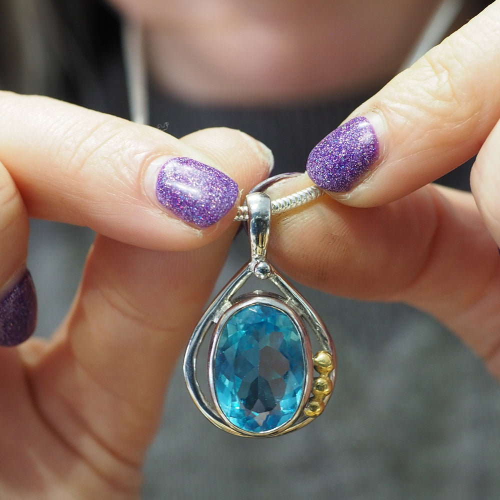 Close-up of hands holding a necklace with a pendant. The Poppy Pendant Small in various gemstones by Gallardo & Blaine Designs features a large, oval-cut blue gemstone set in a sterling silver teardrop-shaped frame adorned with three small gold accents. The person has glittery purple nail polish.