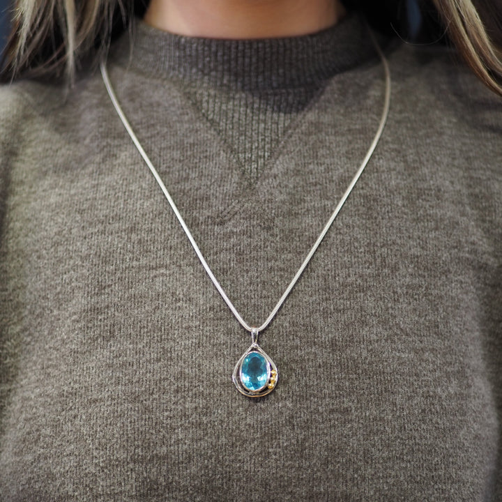 A person wearing a gray sweater has a sterling silver necklace with a teardrop-shaped blue gemstone pendant from Gallardo & Blaine Designs called the Poppy Pendant Small in various gemstones. The background is blurred, focusing on the necklace and sweater.