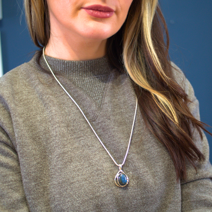 A person wearing a grey sweater is shown from the shoulders up. They have long brown hair with blonde highlights and are wearing a Gallardo & Blaine Designs sterling silver necklace with a large blue Poppy Pendant Small in various gemstones. The background is blurred, focusing attention on the captivating necklace.