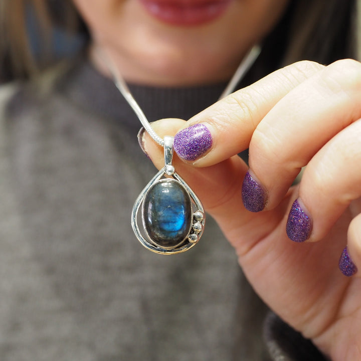 A person with purple glitter nail polish holds a teardrop-shaped Poppy Pendant Small in various gemstones by Gallardo & Blaine Designs encased in a sterling silver setting on a necklace chain, close to the camera. The person is wearing a grey sweater.