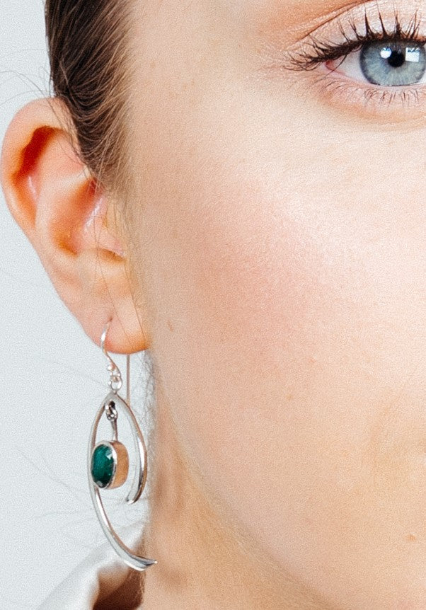 Close-up shot of a person wearing minimalist elegance with a pair of Primrose Earrings in various gemstones by Gallardo & Blaine Designs. The sterling silver earrings feature an oval green gemstone and modern, artistic design with curved silver elements. The person's ear and part of their face are visible, showcasing their fair skin and light eye.