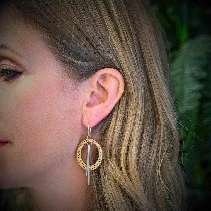 A close-up image of the side of a woman's face, displaying her blonde hair and a unique earring. The Art Deco-style *Gallardo & Blaine Designs Rivera Earrings* are a circular design made of a perforated light-colored material, with a straight metallic rod hanging in the center. The background is green foliage.