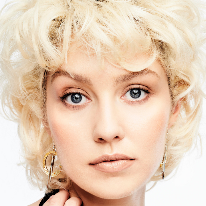 Close-up portrait of a person with short, curly blonde hair, blue eyes, and a neutral facial expression. They are wearing Gallardo & Blaine Designs Rivera Earrings with a circular design and a black outfit, against a plain white background.