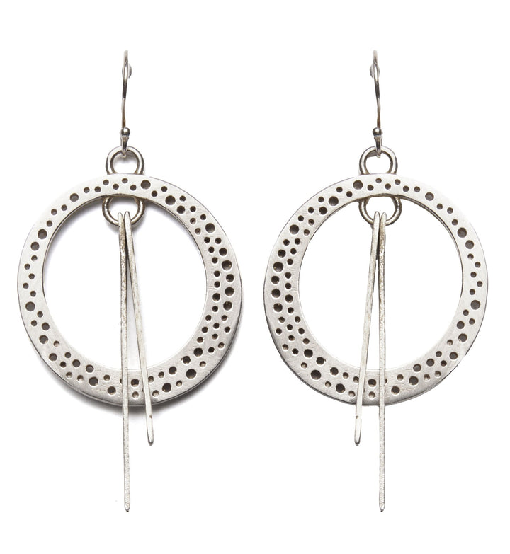 A pair of Rivera Earrings by Gallardo & Blaine Designs featuring large, circular hoops with Art Deco-inspired perforated patterns along the edges. Each hoop has two thin, elongated silver pieces hanging from the center. The earrings are suspended from simple hooks.