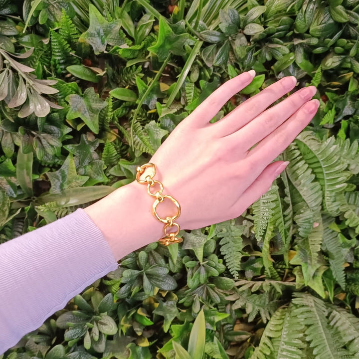 A hand with a light purple sleeve is shown wearing the Sea Honey Bracelet Gold Plated Bronze by Gallardo & Blaine Designs. The background features various lush, green plants and fern leaves.