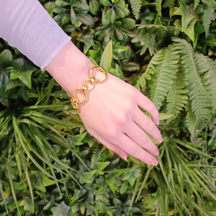 A hand with light skin wearing a **Sea Honey Bracelet Gold Plated Bronze** by **Gallardo & Blaine Designs**, positioned over a background of lush, green leaves and foliage. The sleeve of the wearer is white and slightly translucent.