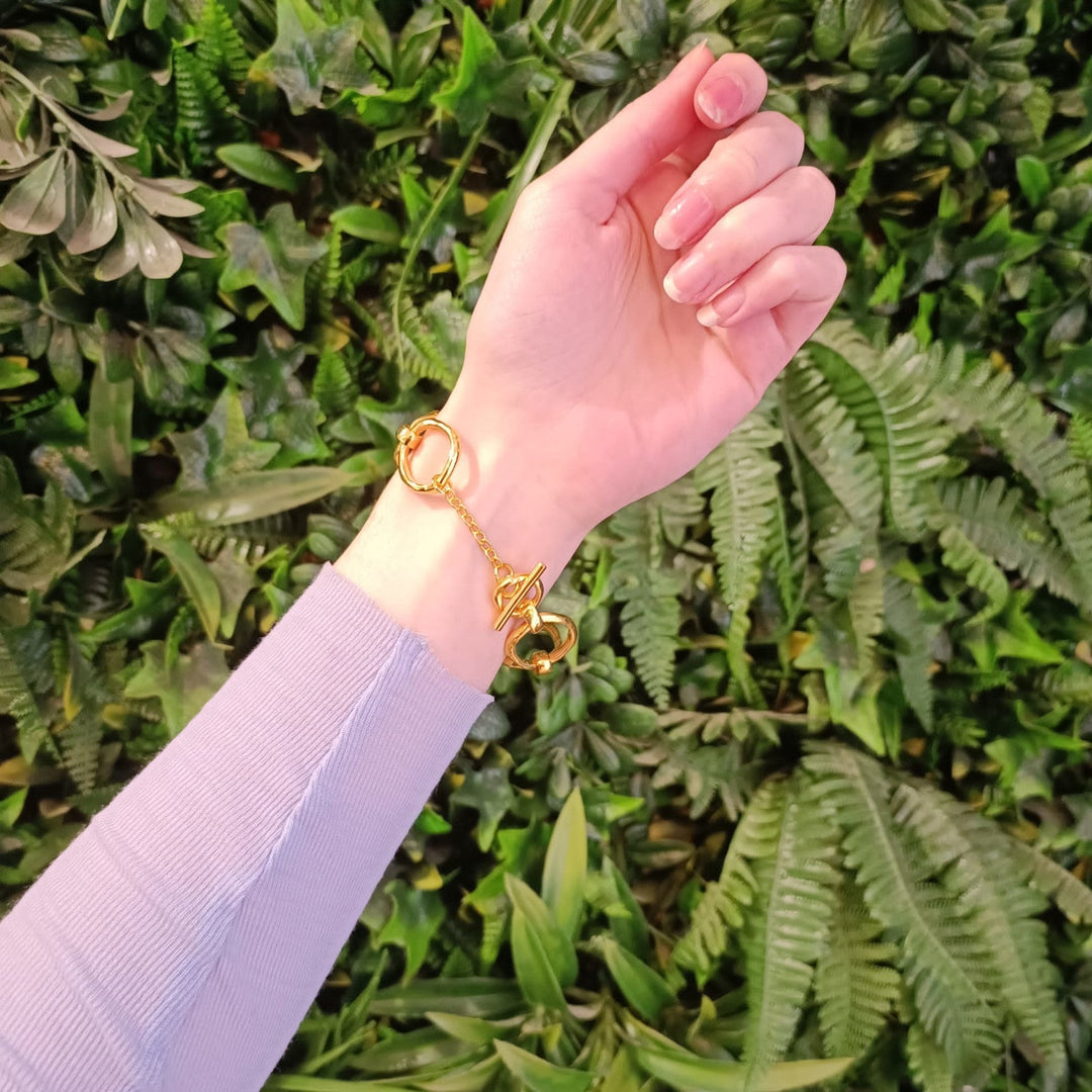 A Sea Honey Bracelet Gold Plated Bronze on a person's wrist against a background of greenery. The gold-colored bracelet glimmers like charm jewelry, and the person is wearing a light purple long-sleeve shirt. The lush greenery consists of various types of leaves and foliage, showcasing Gallardo & Blaine Designs' exquisite craftsmanship.