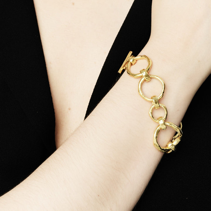 Close-up of a person wearing a black outfit showcasing a bracelet on their wrist. The accessory features large, interconnected circular bronze links with gold plating, adding a stylish touch to the elegant attire. The background is plain, emphasizing the striking Sea Honey Bracelet Gold Plated Bronze by Gallardo & Blaine Designs.