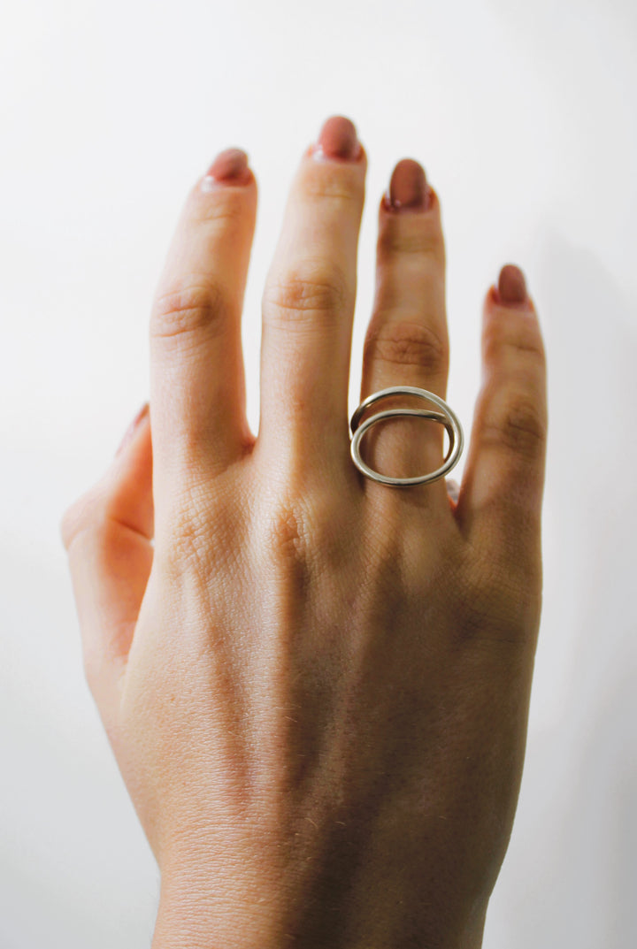 A hand with neatly manicured nails displays the Gallardo & Blaine Designs Sea Honey Ring, a minimalist sterling silver piece with two overlapping ovals, on the ring finger against a plain white background.