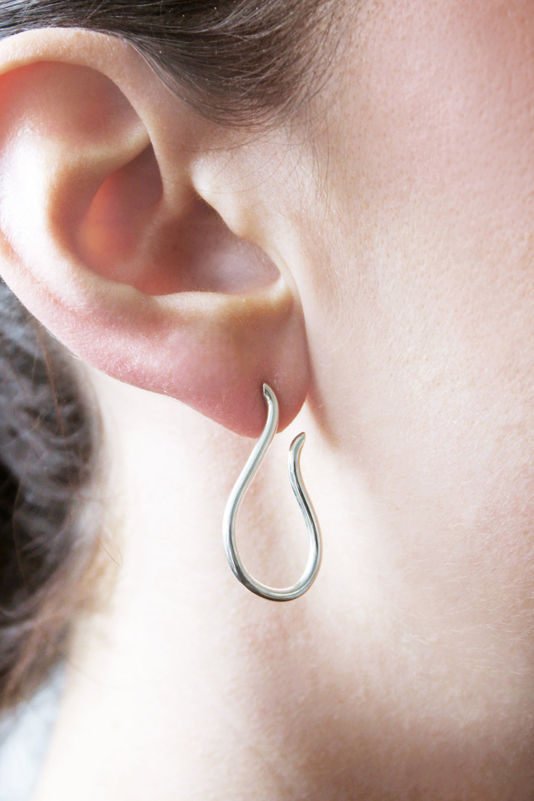 A close-up of a person’s ear wearing Gallardo & Blaine Designs' Sea Honey Studs—a minimalist sterling silver earring with an open end, teardrop hoop shape, and smooth polished finish that highlights its elegant design.