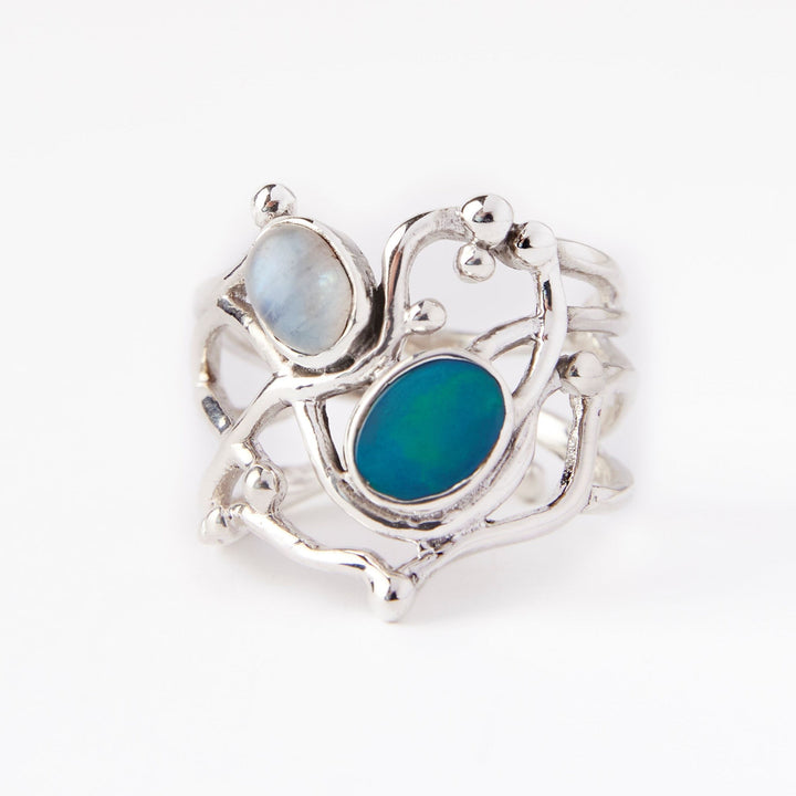 The Seaweed Ring in various gemstones by Gallardo & Blaine Designs in sterling silver features an abstract, wavy design with two oval stones. One stone is a pale, milky color while the other is a vibrant, deep blue-green. The ring's design includes organic, branch-like shapes connecting the stones.