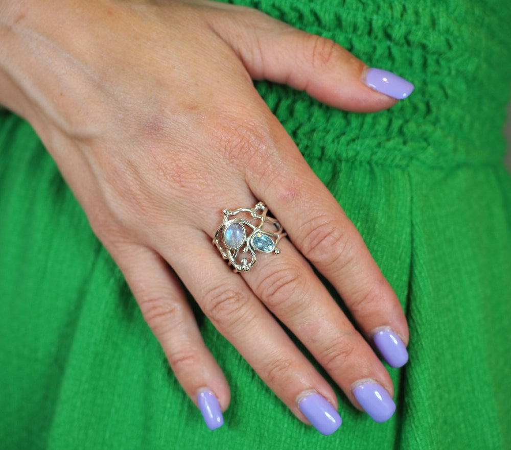 A close-up of a hand with purple-painted nails wearing a gold vermeil Seaweed Ring in various gemstones from Gallardo & Blaine Designs. The hand rests on a green, textured fabric background.