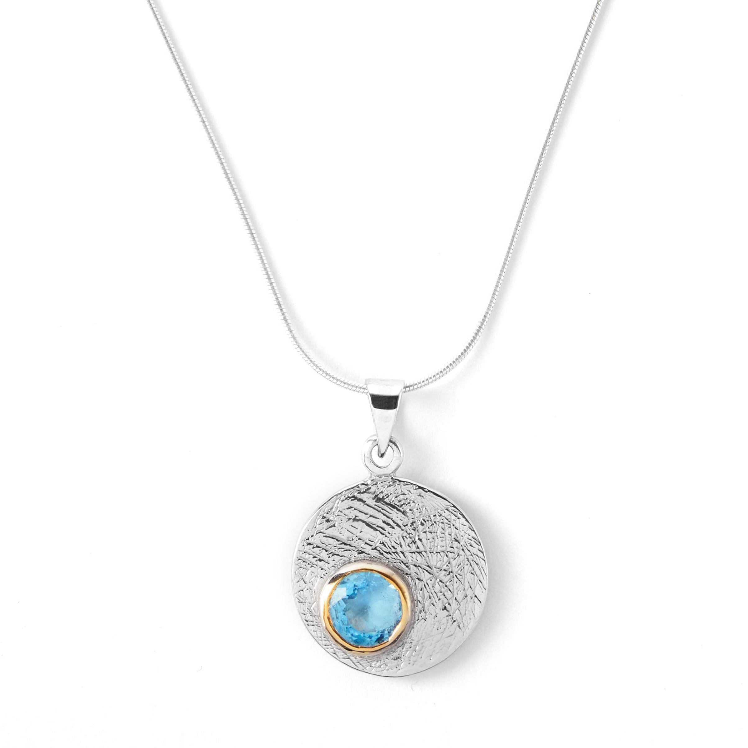 The Selene Necklace Silver - Small in various gemstones by Gallardo & Blaine Designs features a silver pendant with a textured surface and a small, embedded blue stone encircled by a gold bezel. Hanging gracefully from a Sterling silver chain, it combines simplicity and elegance in perfect harmony.