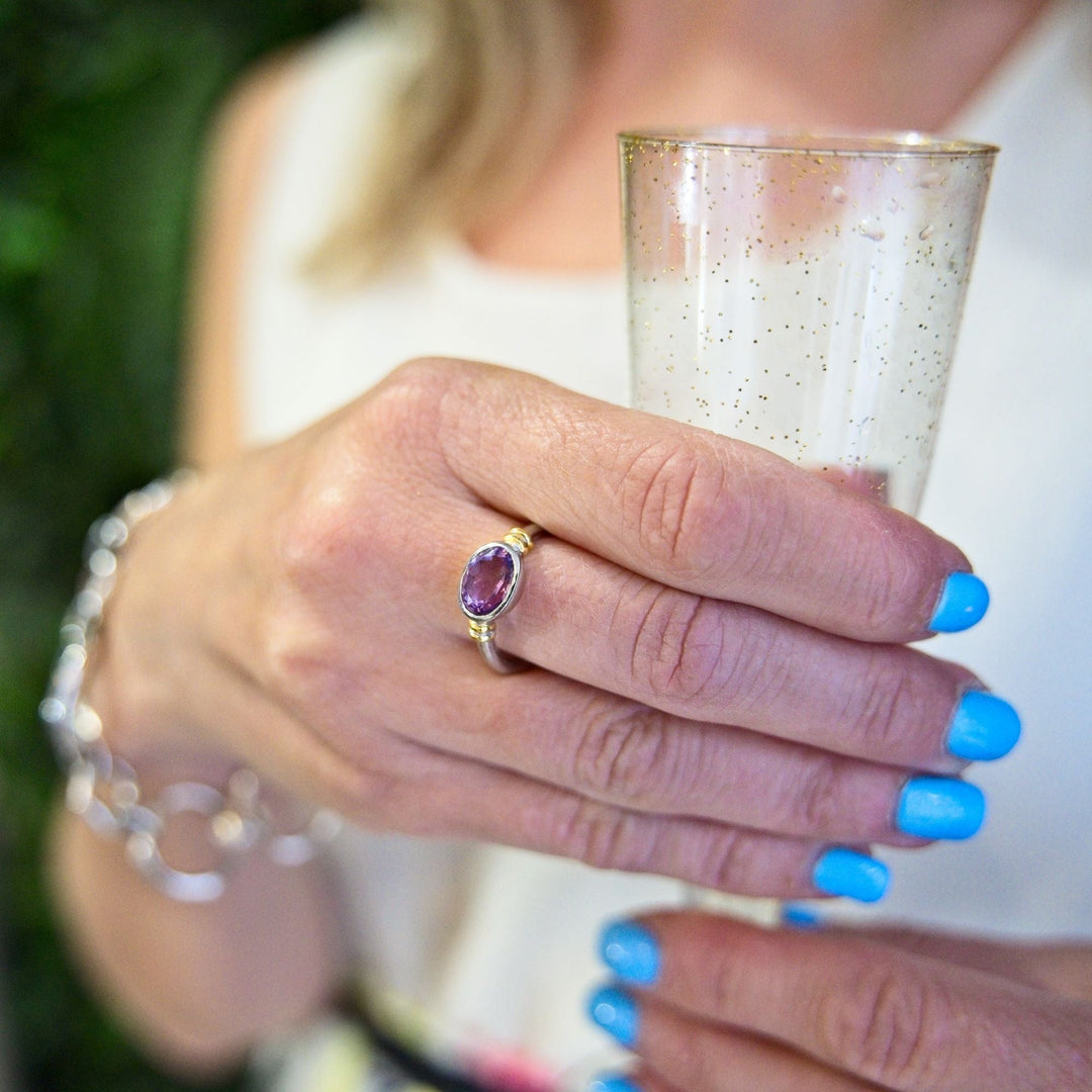 A person with blue manicured nails holds a sparkling drink in a clear, glittery glass. They are wearing the Gallardo & Blaine Designs Senna Ring with a purple gemstone and a silver bracelet. The person is dressed in light-colored clothing.