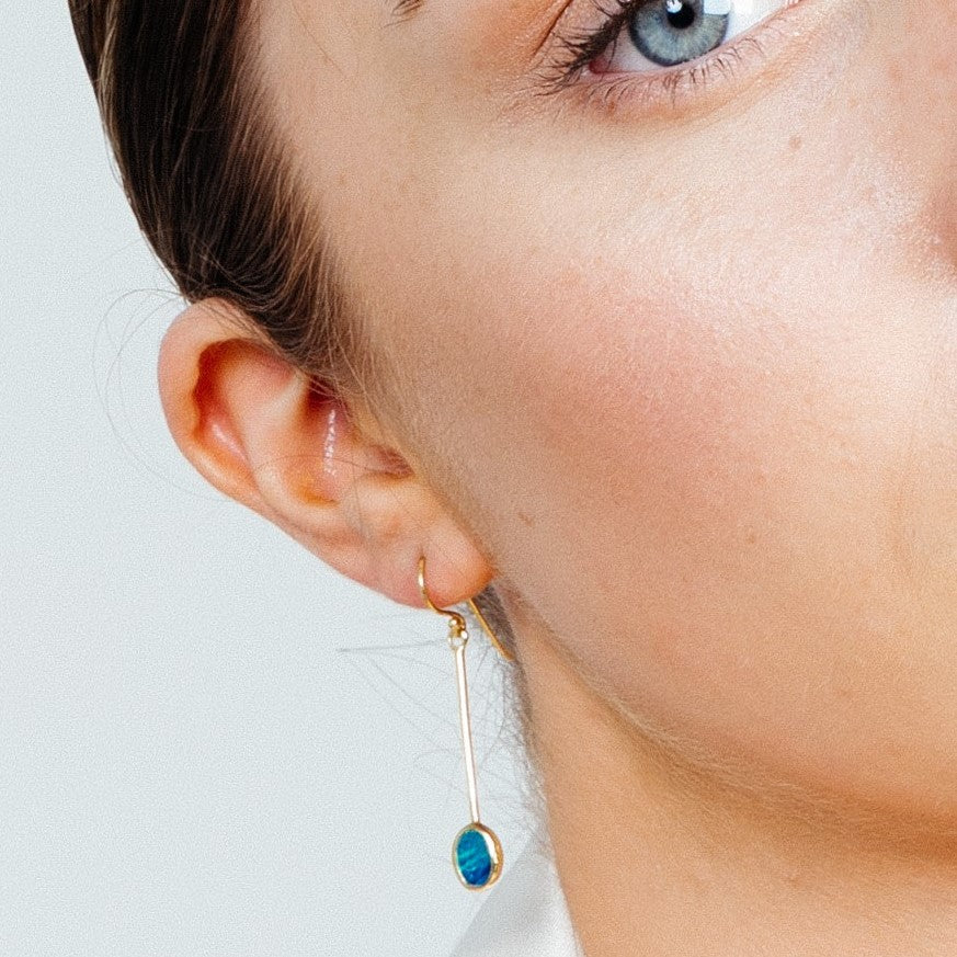 Close-up of a person’s face showing a blue eye, part of their cheek, and ear. They are wearing Sequola Earrings in various gemstones from Gallardo & Blaine Designs: long, slender sterling silver dangle earrings with a blue circular gem. The person's skin is fair, and their dark hair is pulled back. The background is plain and light-colored.