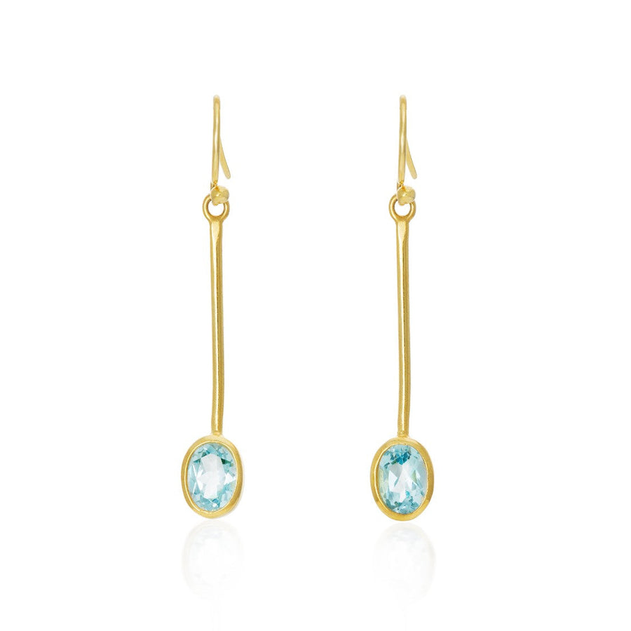 The Sequola Earrings in various gemstones by Gallardo & Blaine Designs are minimalist gold drop earrings featuring thin, elongated bars that hang vertically, each ending with an oval-cut, pale blue gemstone. These sleek and elegant dangle earrings have a simple hook closure at the top, highlighting the delicate stones.