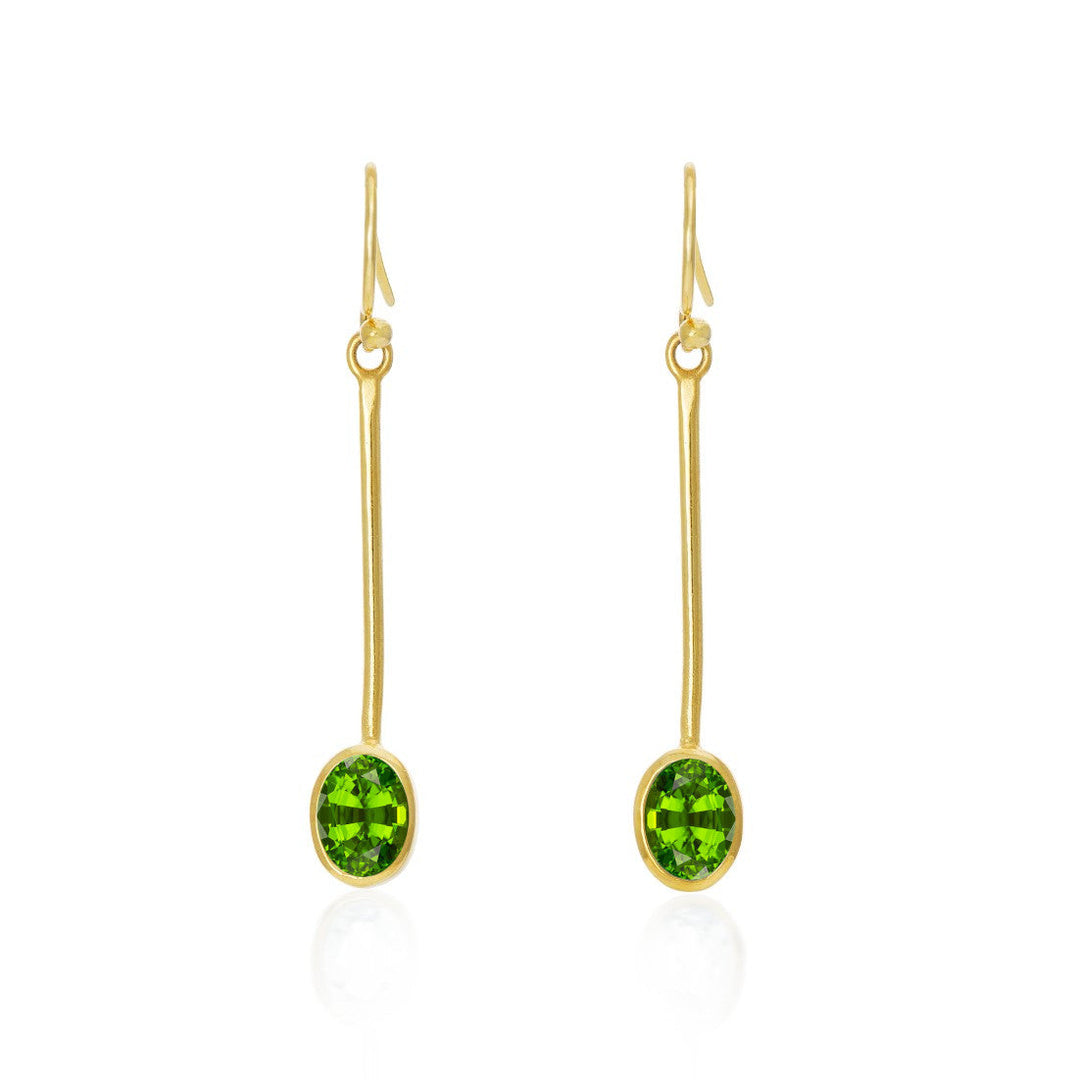 Gallardo & Blaine Designs Sequola Earrings in various gemstones: These gold drop earrings feature elongated hooks and long stems that elegantly end with oval green gemstones. Simple yet sophisticated, they showcase the bright green stones as the focal point against a white background, highlighting their beauty.