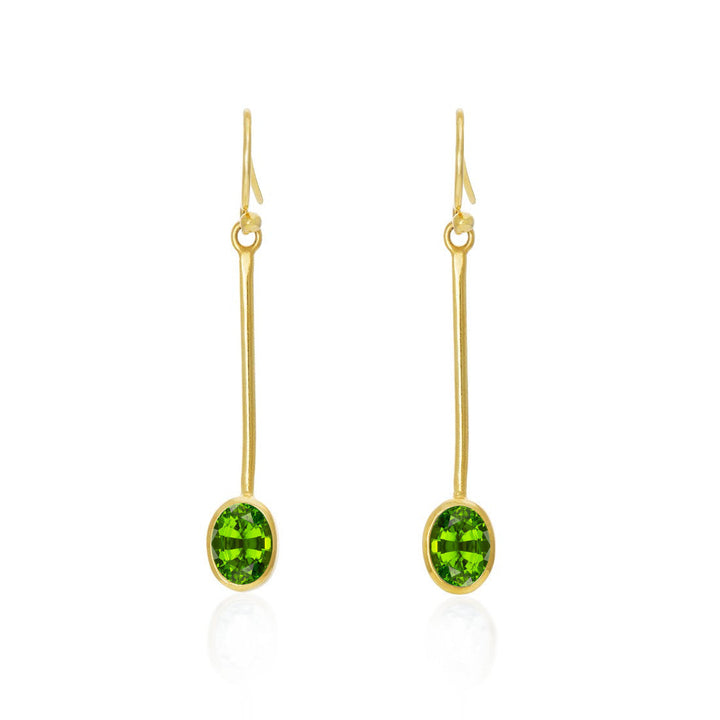 Gallardo & Blaine Designs Sequola Earrings in various gemstones: These gold drop earrings feature elongated hooks and long stems that elegantly end with oval green gemstones. Simple yet sophisticated, they showcase the bright green stones as the focal point against a white background, highlighting their beauty.