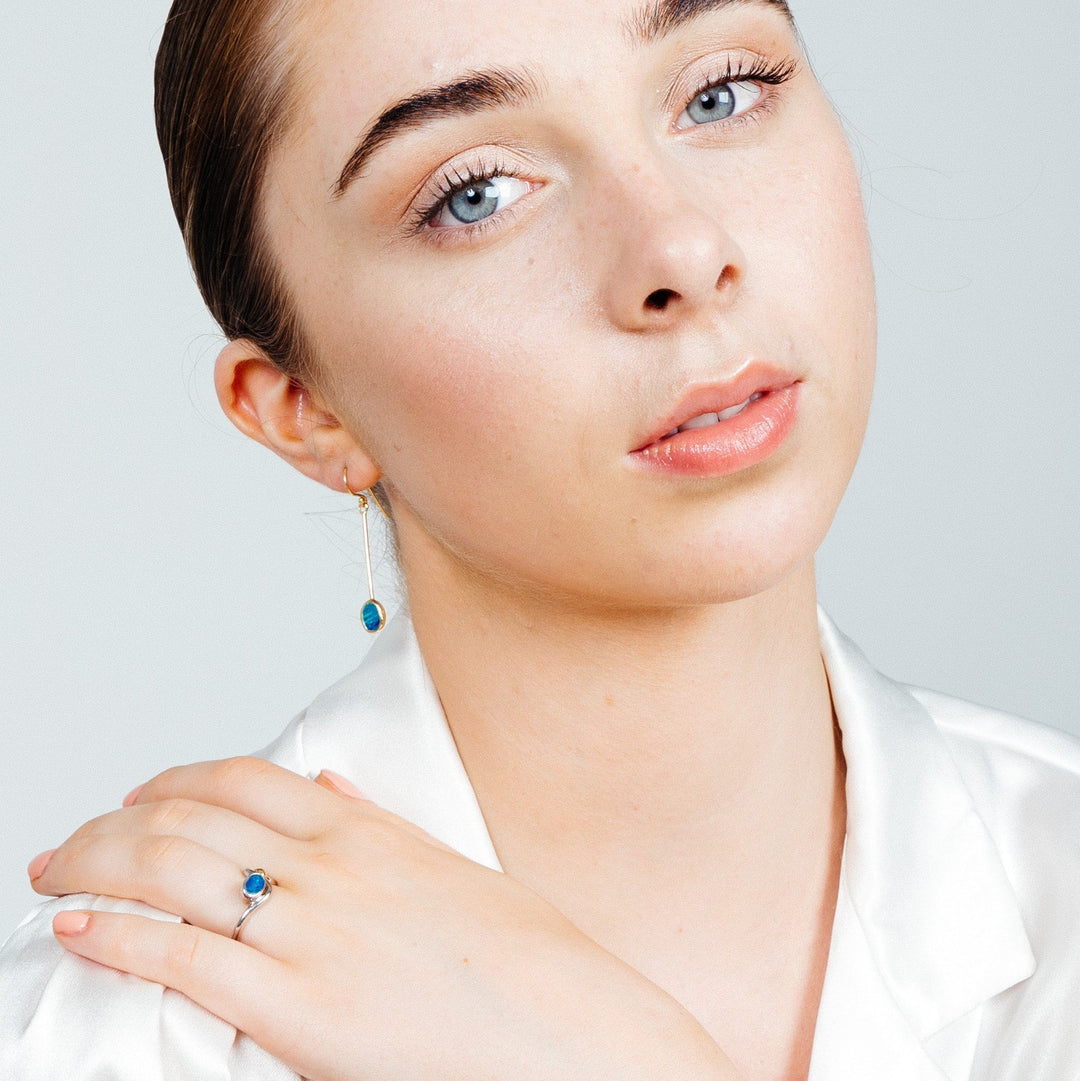 A person with light skin, light blue eyes, and dark hair pulled back is wearing a white blouse. They are adorned with Sequola Earrings in various gemstones by Gallardo & Blaine Designs and a ring, both featuring blue gemstones. The person gazes softly at the camera, resting one hand on their shoulder.