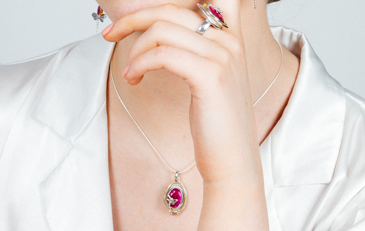 A person wearing a white shirt is adorned with jewelry, including a silver ring with a red gemstone on their middle finger, a Lagoon Pendant Small in various gemstones by Gallardo & Blaine Designs featuring a matching red gemstone, and silver earrings. The person's hand is partially covering their mouth.