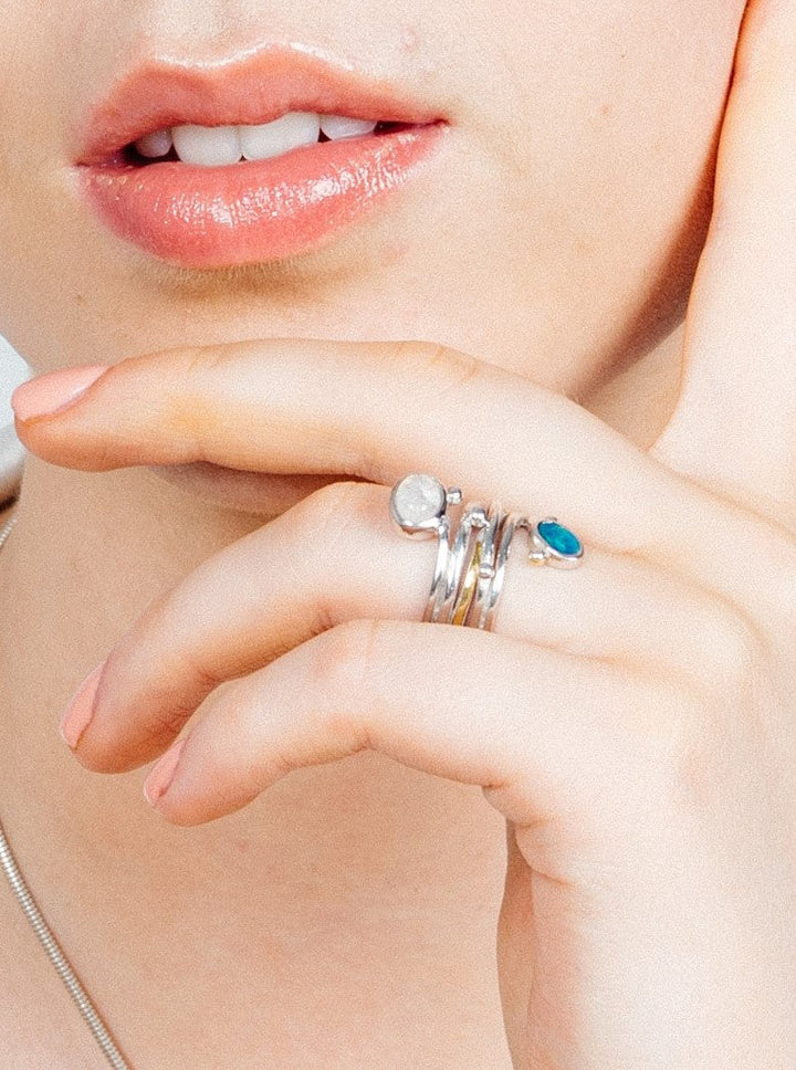 Close-up of a person's lips and hand. The person is wearing four silver rings on their index finger, one with a white stone and another with a unique turquoise design. A Snake Ring in Silver Gold & various gemstones by Gallardo & Blaine Designs graces their middle finger. Their lips are slightly parted, revealing their teeth. The hand is near their face, touching their chin.