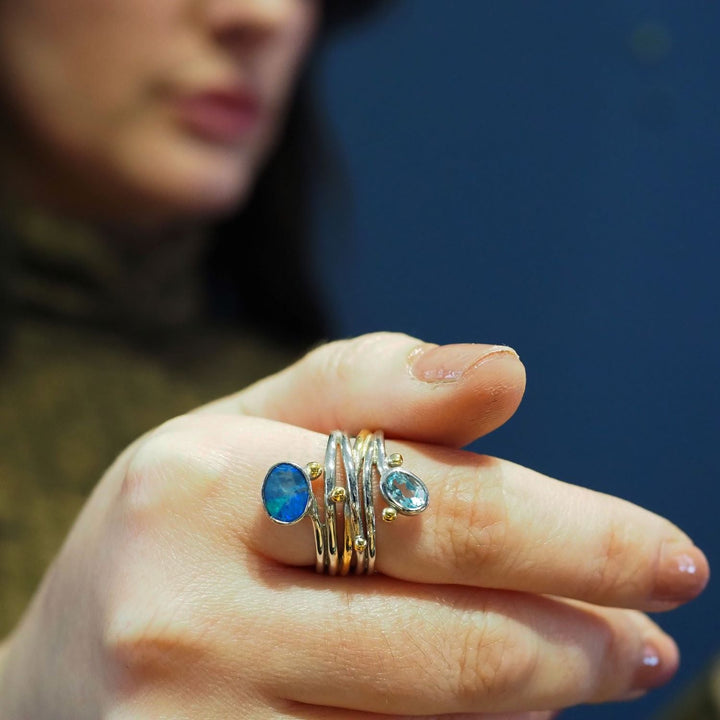 A close-up of a hand wearing several adjustable silver & gold rings with blue gemstones, including a striking Snake Ring in Silver Gold & various gemstones by Gallardo & Blaine Designs. The person’s face is blurred and partially visible in the background against a dark blue backdrop.