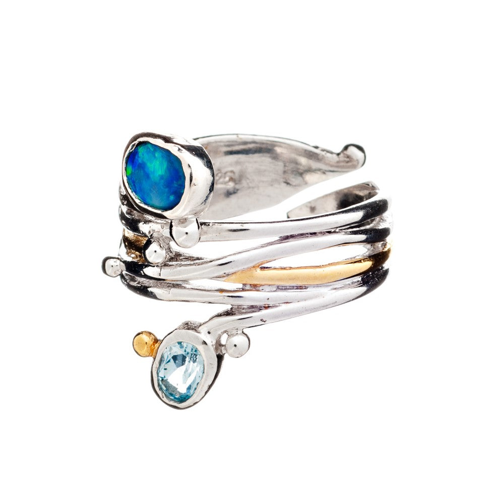 A unique design, the Snake Ring in Silver Gold & various gemstones by Gallardo & Blaine Designs features two gemstones: one round and blue, the other oval and iridescent with green and blue hues. The metal elegantly wraps around the finger adorned with small silver and gold accents.