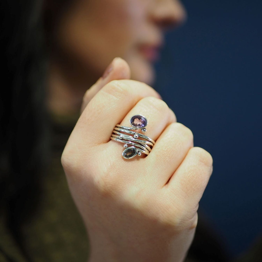 Close-up of a person's hand displaying silver rings with gemstones of varying colors and sizes on their fingers. Among them, a distinctive unique design featuring an adjustable Gallardo & Blaine Designs Snake Ring in Silver Gold & various gemstones stands out. The background is a blurred, dark blue, and the person's face is partially visible but out of focus.