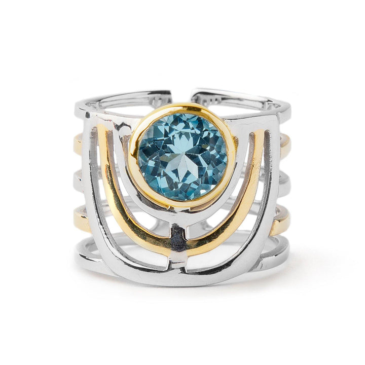 A bold jewellery design, the Solar Ring in various gemstones by Gallardo & Blaine Designs showcases a geometric pattern with a prominent blue gemstone at its heart. The open band structure is accented with gold, artistically framing the gemstone that's encased in a stunning gold setting.