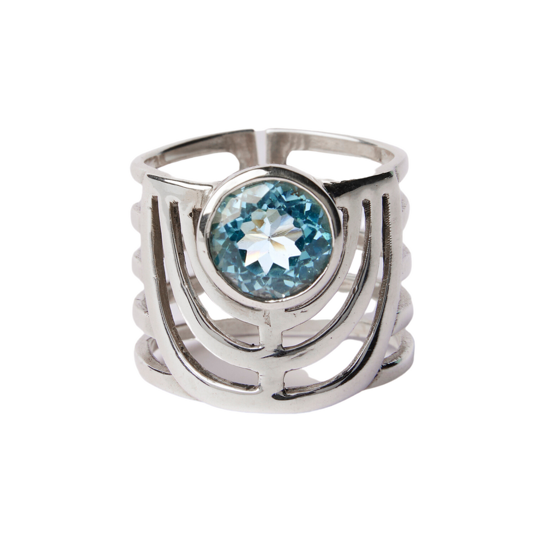 A bold jewellery design, this Solar Ring in various gemstones by Gallardo & Blaine Designs features a unique, openwork pattern with three parallel bands that converge and diverge around a centrally set, round blue gemstone. The intricate setting highlights the sparkling blue stone, making it the focal point of this chunky gemstone ring.