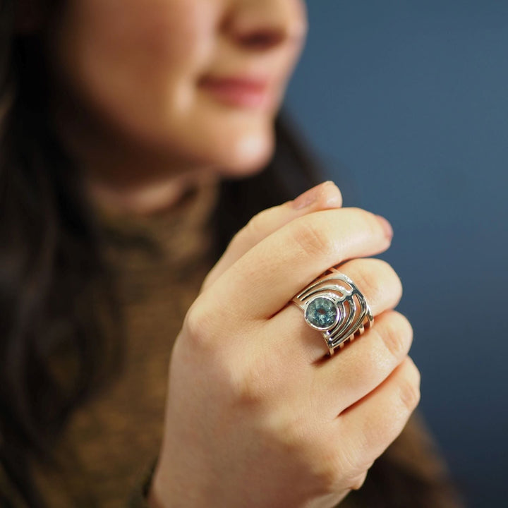 A person holding their hand near their face, wearing a bold jewellery design featuring the Gallardo & Blaine Designs Solar Ring in various gemstones with multiple bands and a round blue gemstone. The background is blurred, making the chunky gemstone ring the focal point of the image. The person is partially visible, with a blurred face and long hair.